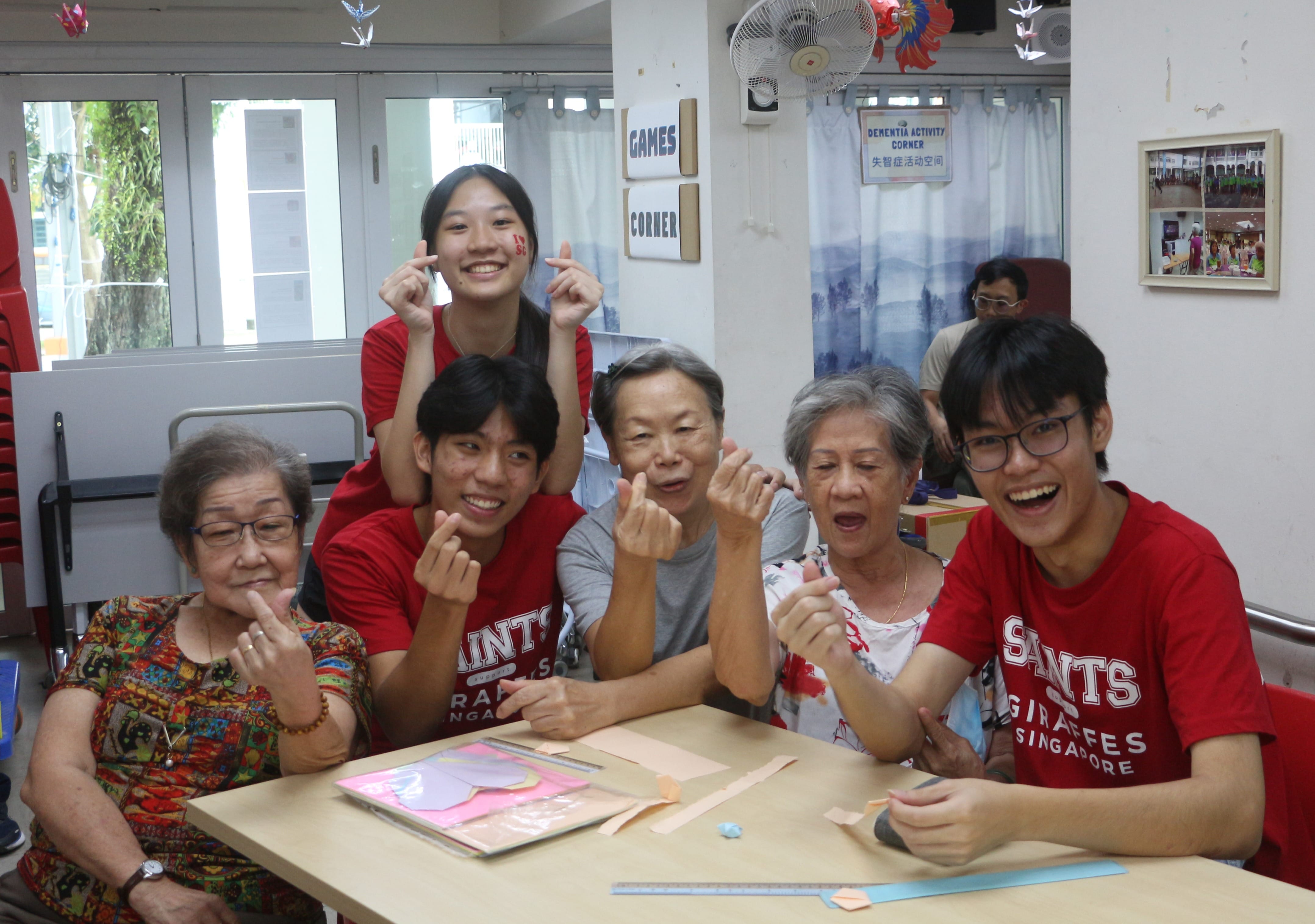 Student volunteers in red shirts doing origami crafts with elderly residents around a table, with everyone making finger-heart hand gestures.